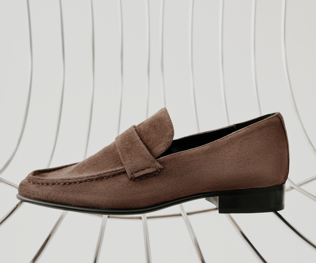 A single brown suede penny loafer with a low black block heel, displayed on a white wire chair against a white background with vertical line patterns.