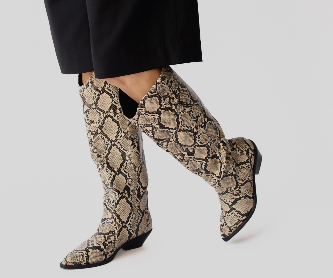 A pair of knee-high pointed-toe boots with a beige and brown snakeskin pattern, shown on a model wearing black trousers against a light gray background.